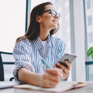 Eine junge Frau in einem gepunkteten Hemd sitzt lächelnd an einem Arbeitsplatz mit Blick aus dem Fenster und hält ein Smartphone in der Hand. Dieses Bild könnte einen Moment des Innehaltens während der Arbeit darstellen, vielleicht während sie eine erfreuliche Nachricht liest oder eine kleine Pause macht.