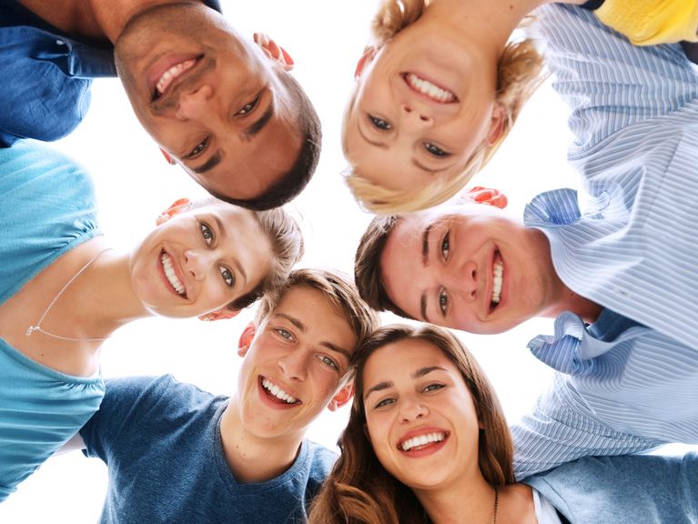 Ein fröhlicher Freundeskreis bildet einen Kreis und blickt von oben in die Kamera, wobei ihre Gesichter ein Bild der Verbundenheit und Freude formen. Die verschiedenen Blautöne ihrer Kleidung kontrastieren schön mit dem hellen Hintergrund, was ein Gefühl von Leichtigkeit und Glück vermittelt.