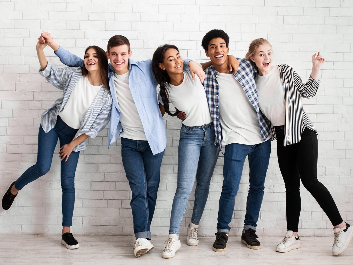 Fünf fröhliche Freunde posieren für ein Foto, während sie in einer Reihe stehen und ausgelassen gestikulieren, gegen eine weiße Ziegelwand gelehnt.