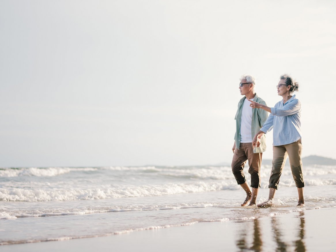 Ein älteres Paar spaziert entspannt am Strand, Hand in Hand, mit dem Blick auf das Meer gerichtet. Die Szene strahlt Ruhe und Verbundenheit aus und zeigt, wie sie gemeinsam den Moment genießen.