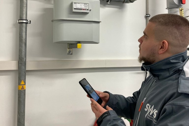 Ein Zählerableser der Stadtwerke Stralsund prüft aufmerksam einen Gaszähler und hält ein mobiles Erfassungsgerät in der Hand in einem technischen Raum.