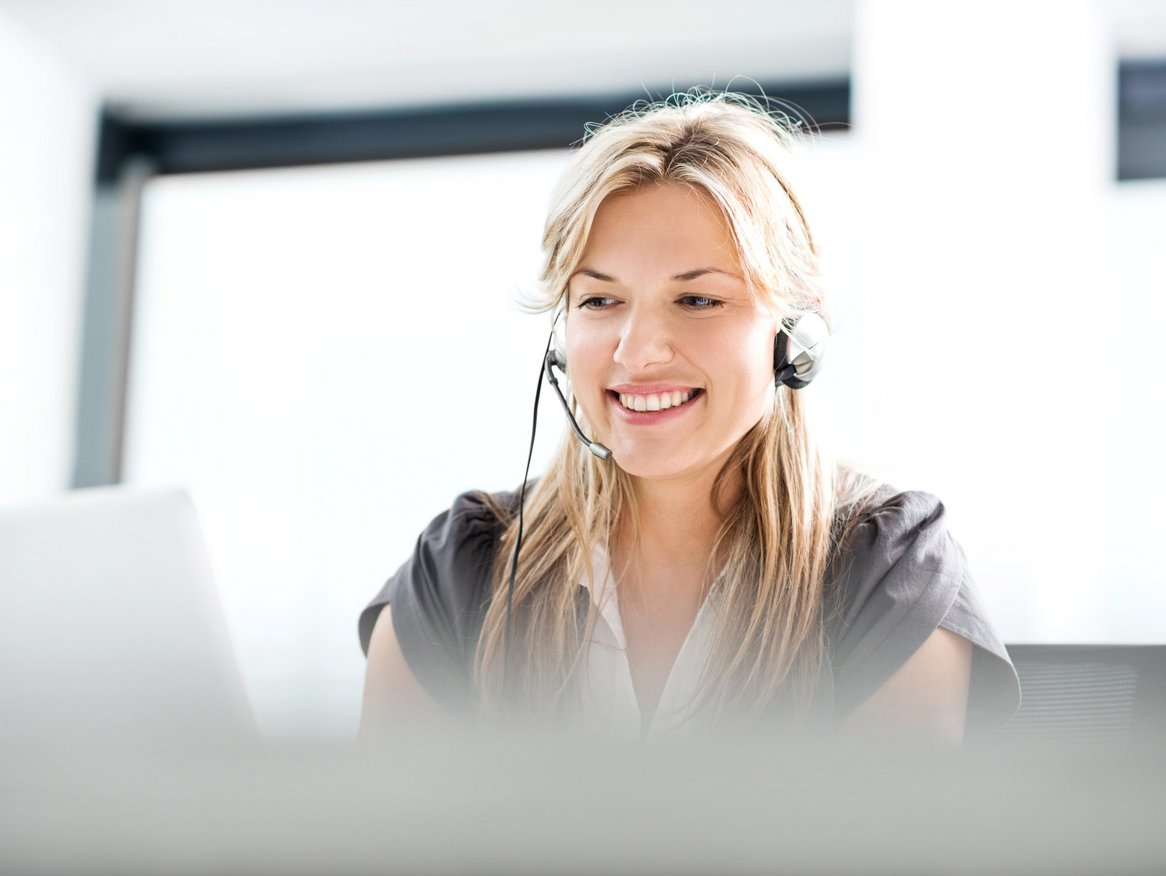 Eine lächelnde Frau mit Headset sitzt vor einem Laptop, anscheinend in einem hellen Büro oder zu Hause, während sie ein freundliches Kundengespräch führt oder an einem virtuellen Meeting teilnimmt.