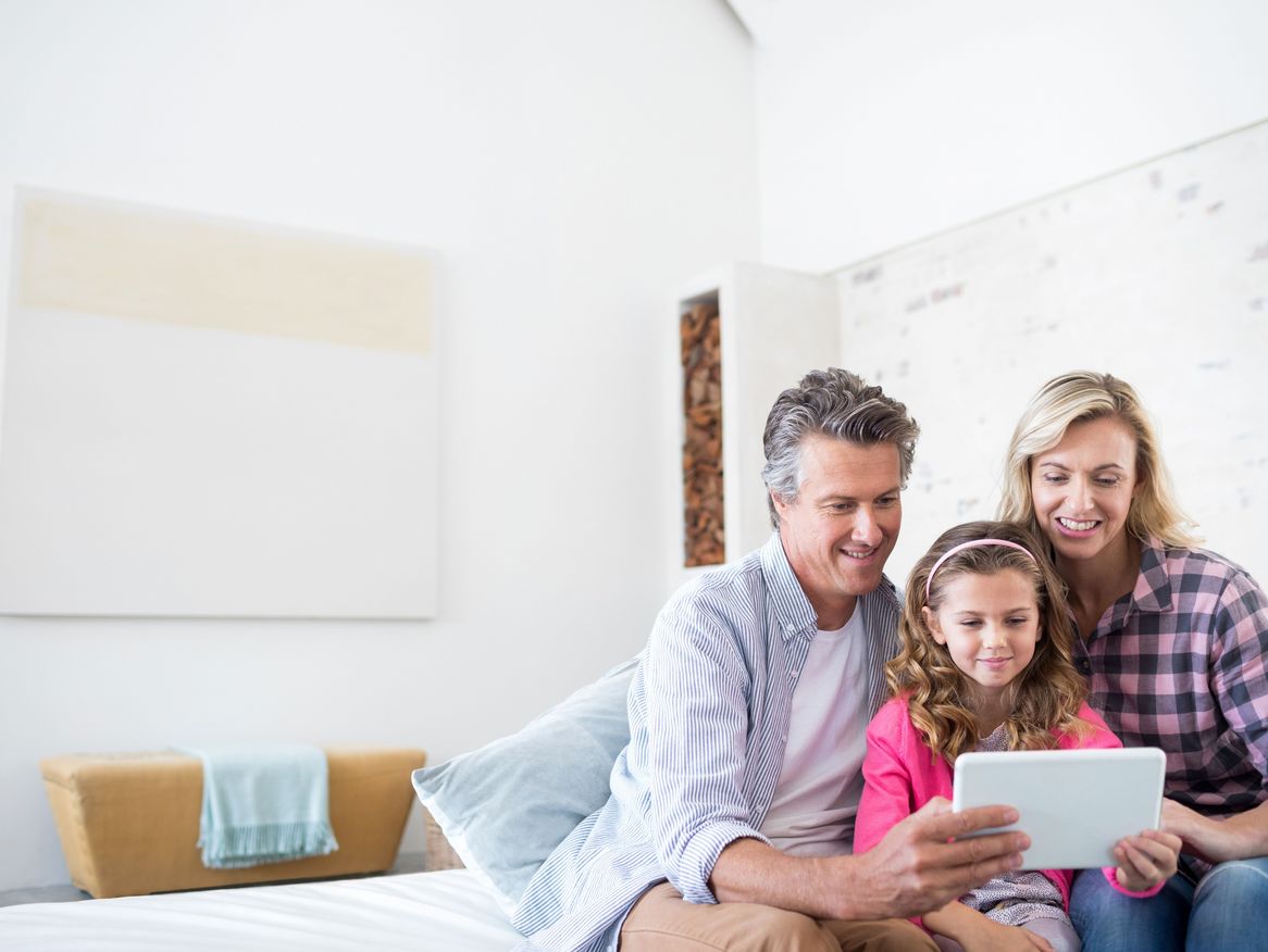 Eine Familie mit einem Kind sitzt gemeinsam auf dem Bett und betrachtet ein Tablet. Sie lächeln und genießen eine gemütliche Zeit in einem hellen, modern eingerichteten Schlafzimmer.