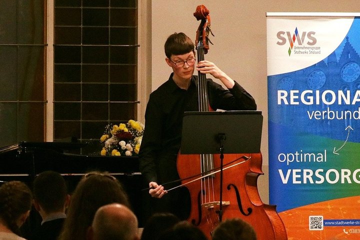 Ein Cellist spielt leidenschaftlich vor einem Publikum, im Hintergrund ist ein Banner mit der Aufschrift "REGIONAL... VERSORGUNG" zu sehen