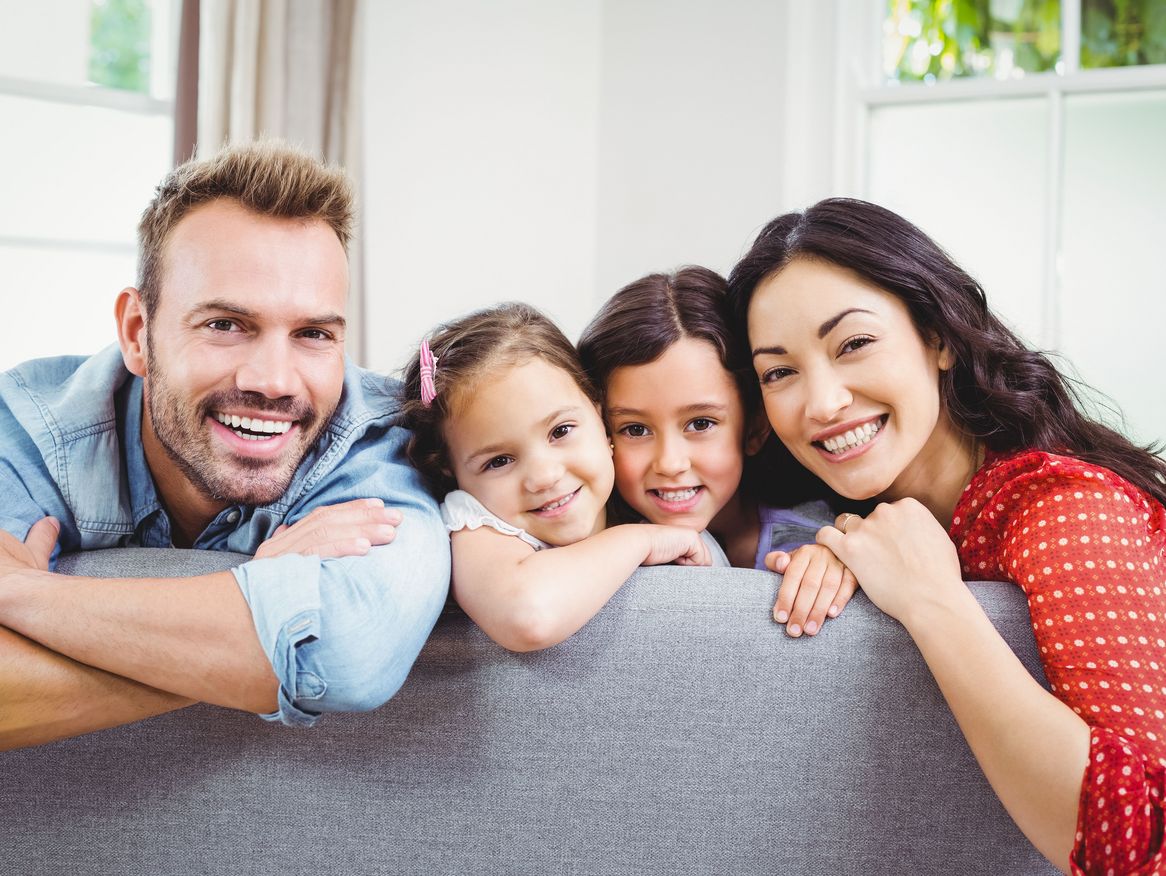 Eine glückliche Familie lächelt in die Kamera, während sie gemütlich auf dem Sofa sitzt, mit einem hellen Wohnzimmer im Hintergrund.