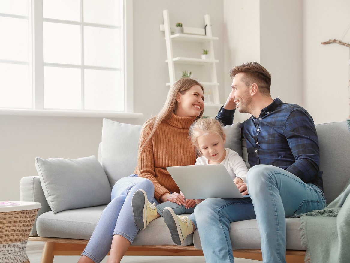 Eine junge Familie sitzt gemütlich auf einem Sofa in einem hellen Wohnzimmer, die Mutter hält ein Kleinkind auf dem Schoß und benutzt einen Laptop, während sie und der Vater lächelnd miteinander interagieren.