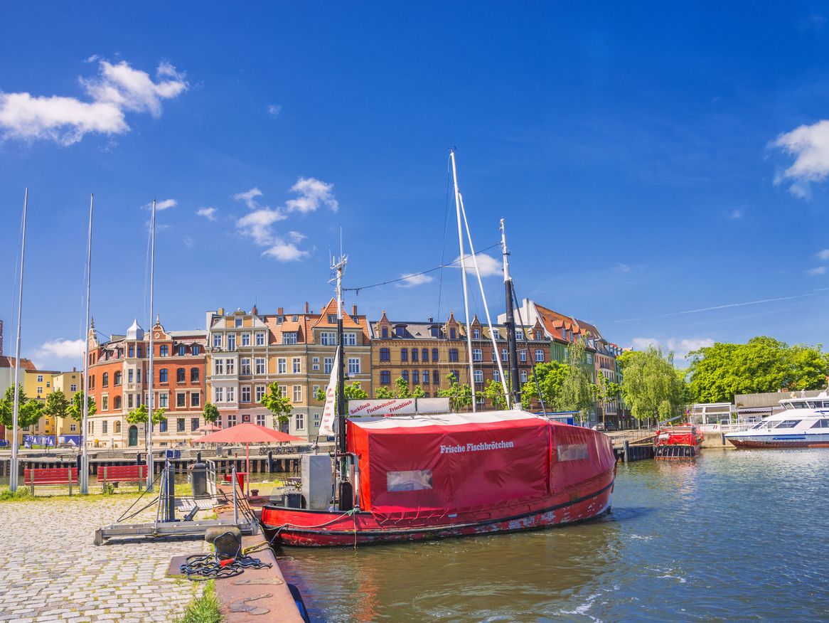 Das Bild zeigt eine malerische Uferpromenade mit einem roten Boot im Vordergrund und historischen Gebäuden im Hintergrund unter einem strahlend blauen Himmel. Die Szene ist charakteristisch für eine belebte Hafenstadt und strahlt eine lebendige, sommerliche Atmosphäre aus.