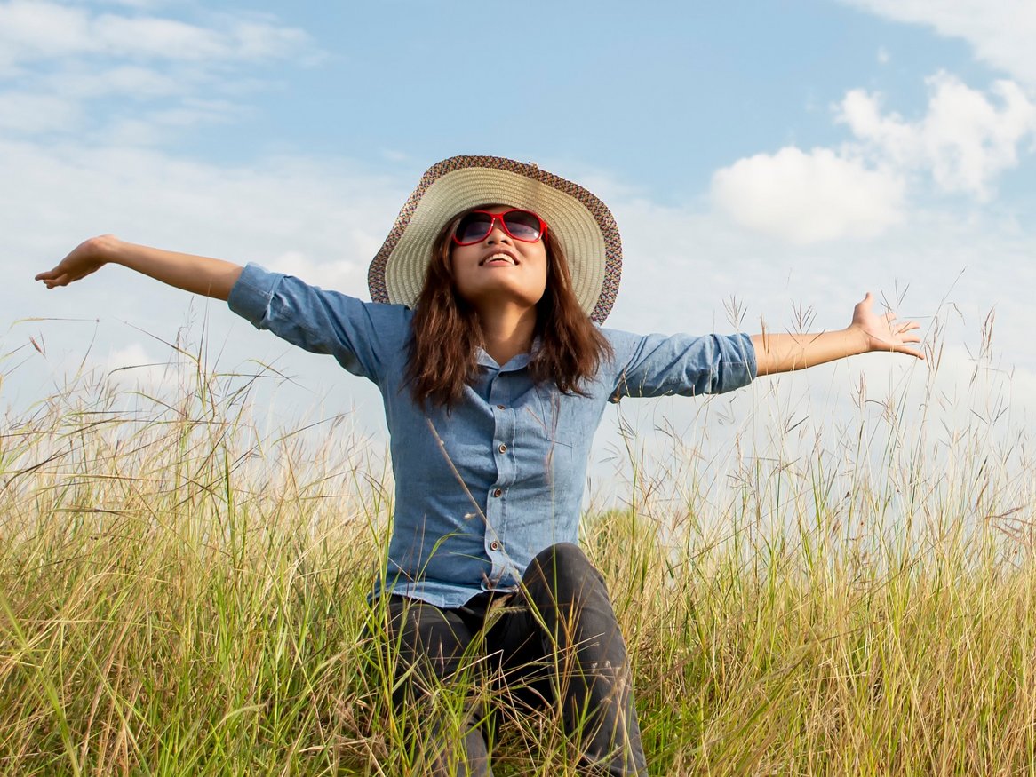 Ein Mädchen im blauen Kleid und mit einem Strohhut genießt die Freiheit, läuft durch ein Feld mit hohen Gräsern und streckt die Arme aus, während sie von der Sonne umgeben ist.