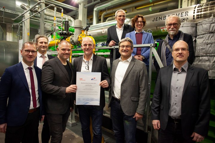 Eine Gruppe von acht Männern, gekleidet in Business-Kleidung, steht stolz in einem Industrieumfeld mit einem großen Maschinenaggregat im Hintergrund.