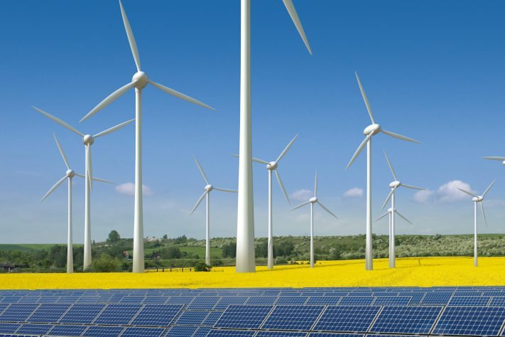Eine Reihe von Windkraftanlagen in einer ländlichen Landschaft mit leuchtend gelbem Rapsfeld im Vordergrund und einem klaren blauen Himmel im Hintergrund, ein Bild für erneuerbare Energien und saubere Stromerzeugung.