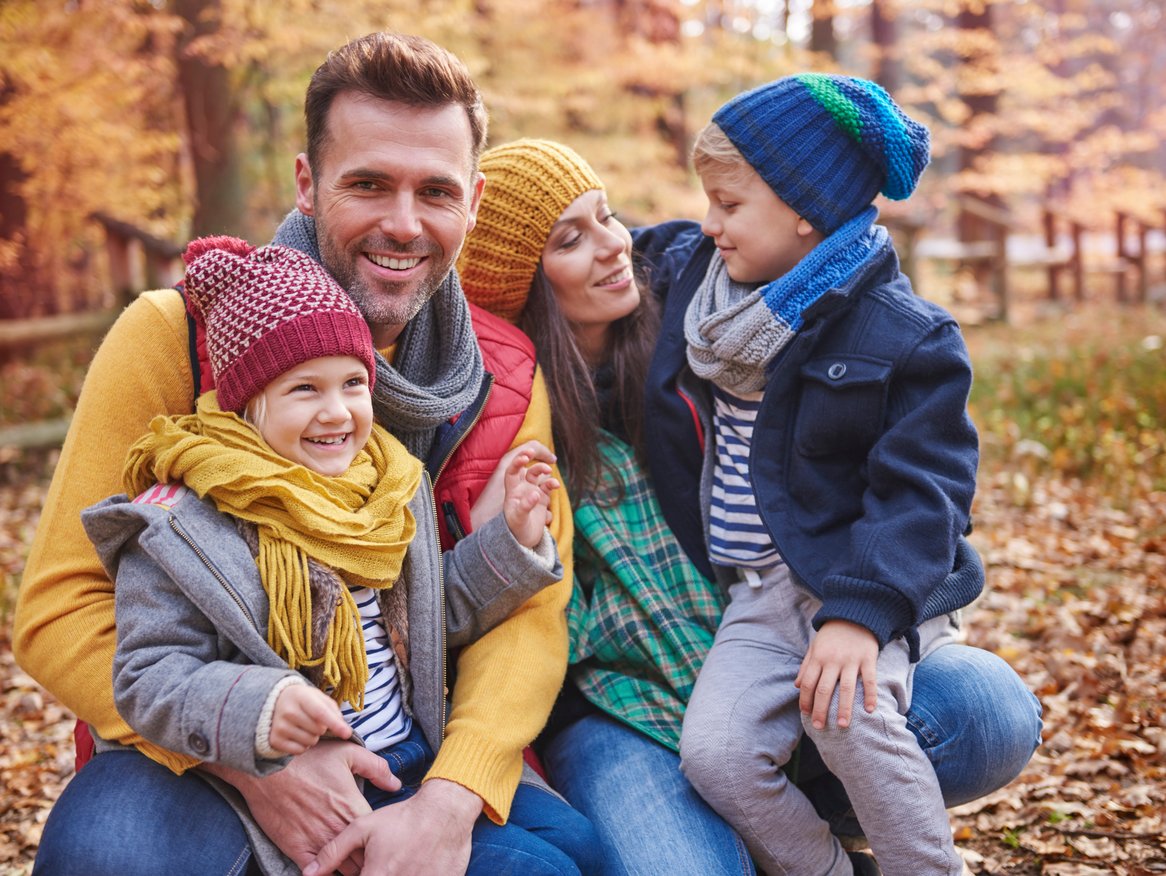 Eine vierköpfige Familie genießt einen sonnigen Herbsttag im Park, wo zwei Kinder mit bunten Strickmützen sich an ihre lächelnden Eltern kuscheln, umgeben von einem Teppich aus goldenen Herbstblättern.