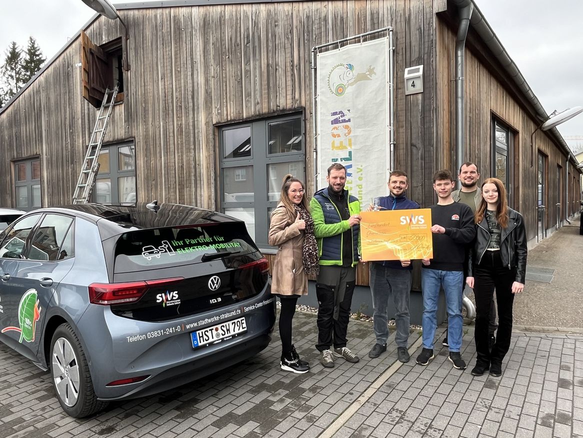 Das Bild zeigt fünf Personen, die stolz neben einem grauen Elektroauto stehen, das mit Werbeaufklebern versehen ist.