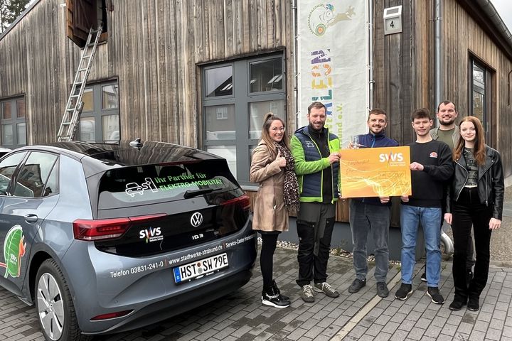 Das Bild zeigt fünf Personen, die stolz neben einem grauen Elektroauto stehen, das mit Werbeaufklebern versehen ist.