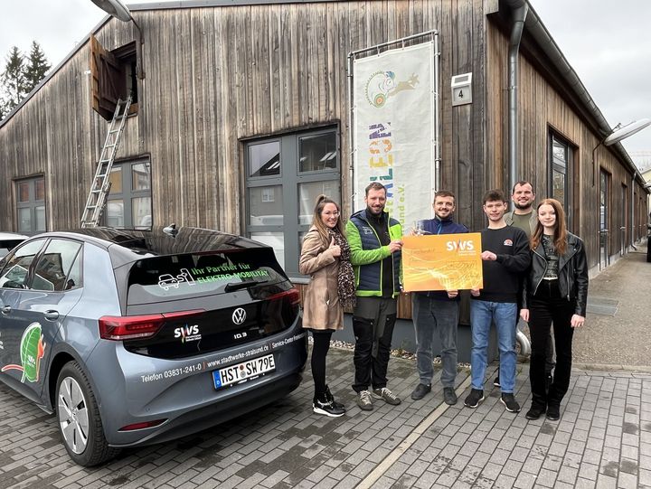 Das Bild zeigt fünf Personen, die stolz neben einem grauen Elektroauto stehen, das mit Werbeaufklebern versehen ist.
