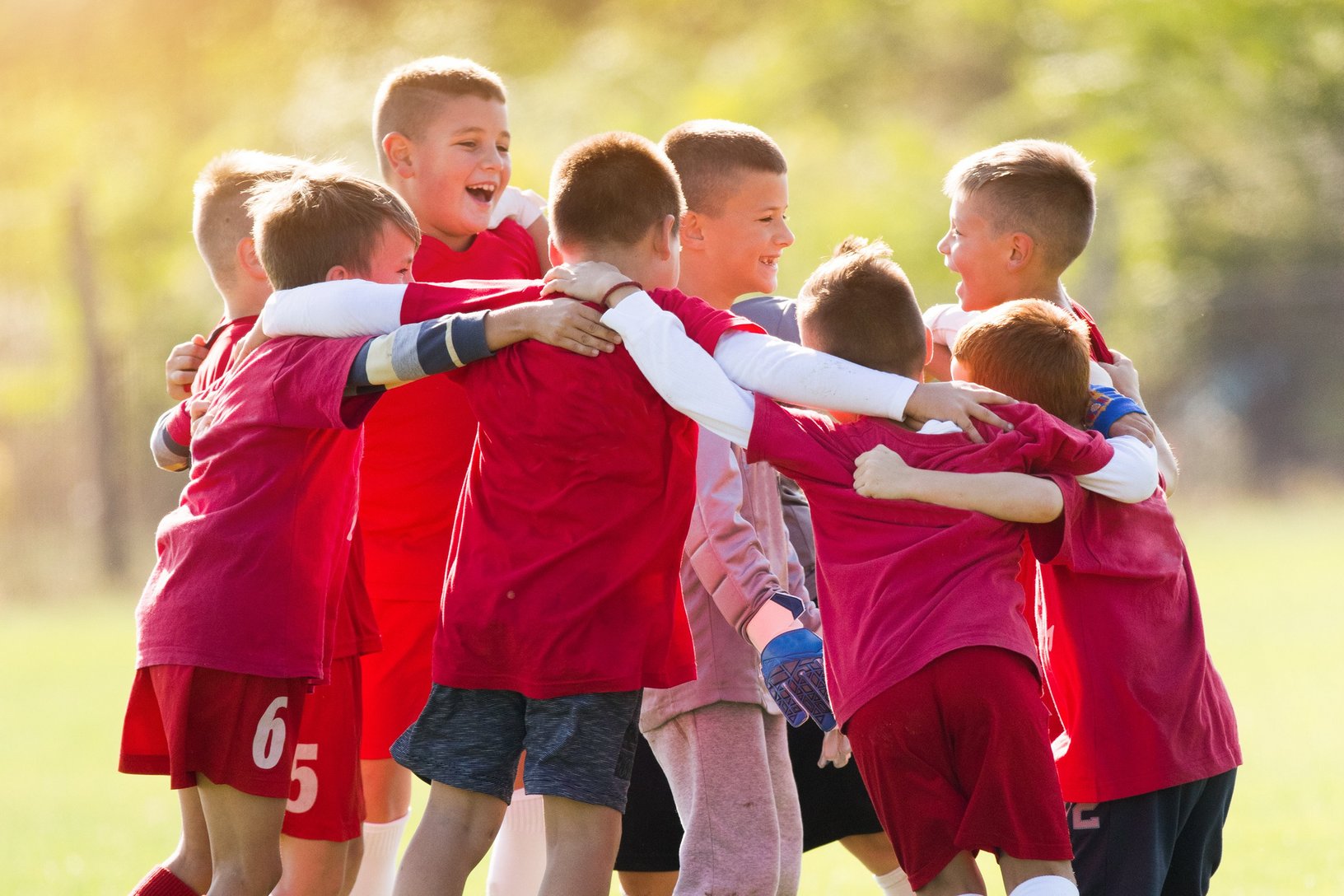 Eine Gruppe von jungen Fußballspielern in roten Trikots bildet einen Kreis und legt die Arme umeinander, ein Ausdruck von Teamgeist und Zusammenhalt vor oder nach einem Spiel. Ein Trainer beobachtet sie im Hintergrund, was die Bedeutung von Führung und Mentorship im Jugendsport unterstreicht.