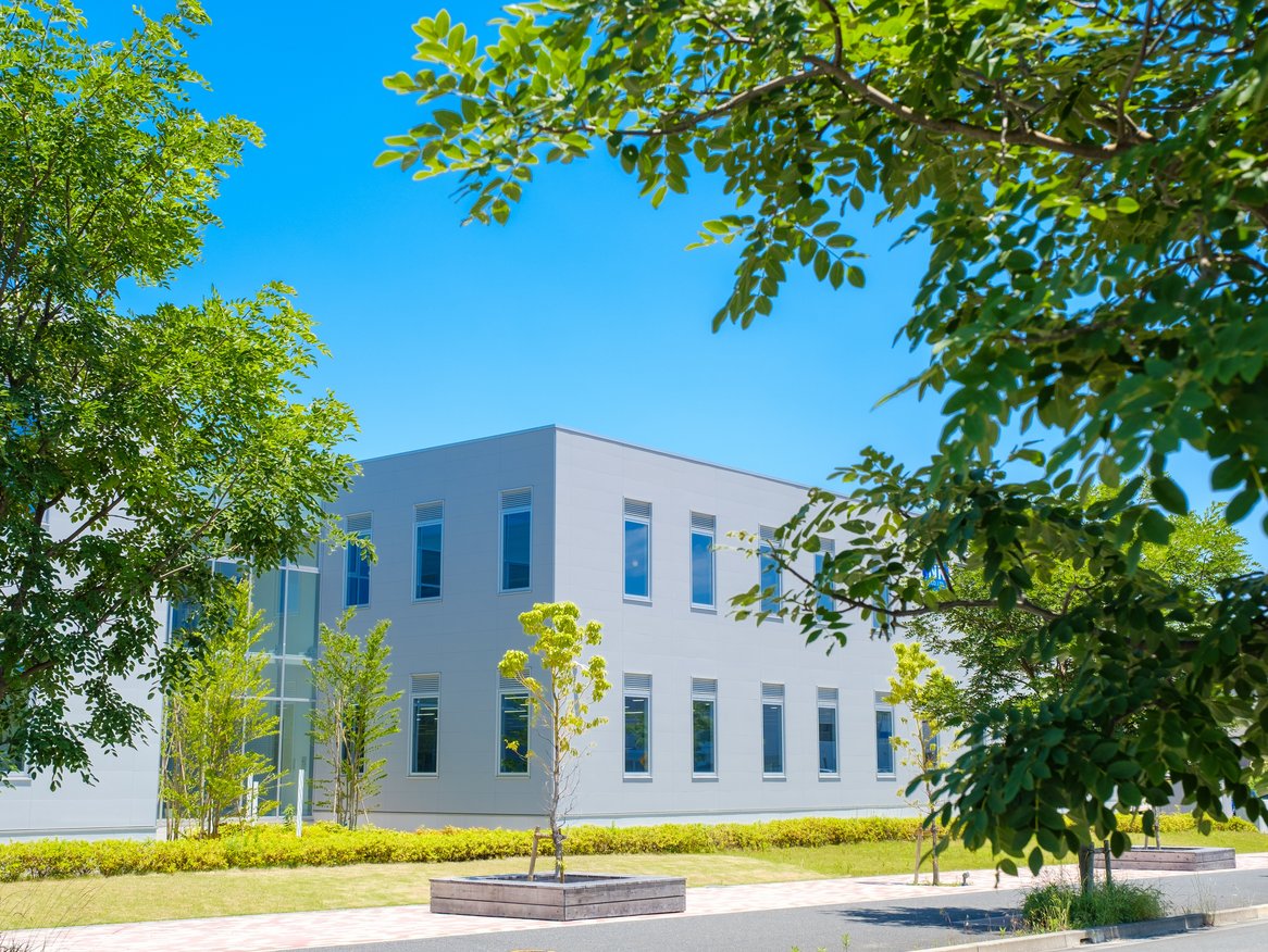 Ein modernes, graues Bürogebäude, eingebettet in eine grüne Landschaft mit reichlich Bäumen unter einem strahlend blauen Himmel an einem sonnigen Tag.