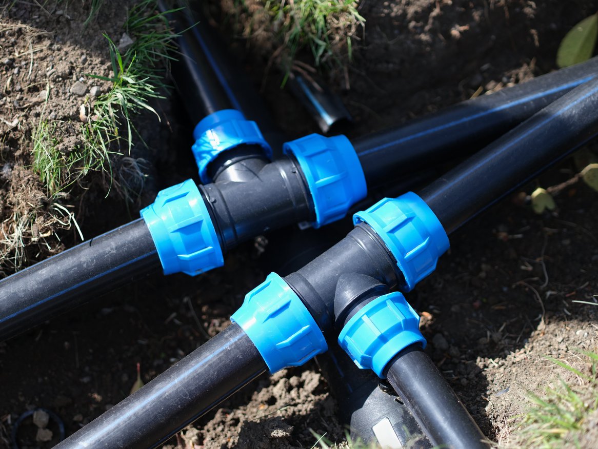 Schwarze Kunststoffrohre mit blauen Verbindungsstücken im Erdreich verlegt, vermutlich für Wasser- oder Bewässerungssysteme.