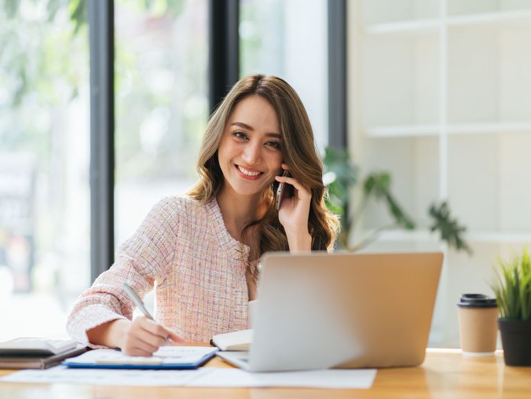 Eine lächelnde Frau am Arbeitsplatz führt ein Telefongespräch und benutzt gleichzeitig einen Laptop, ein Bild von Multitasking und professionellem Arbeitsumfeld.