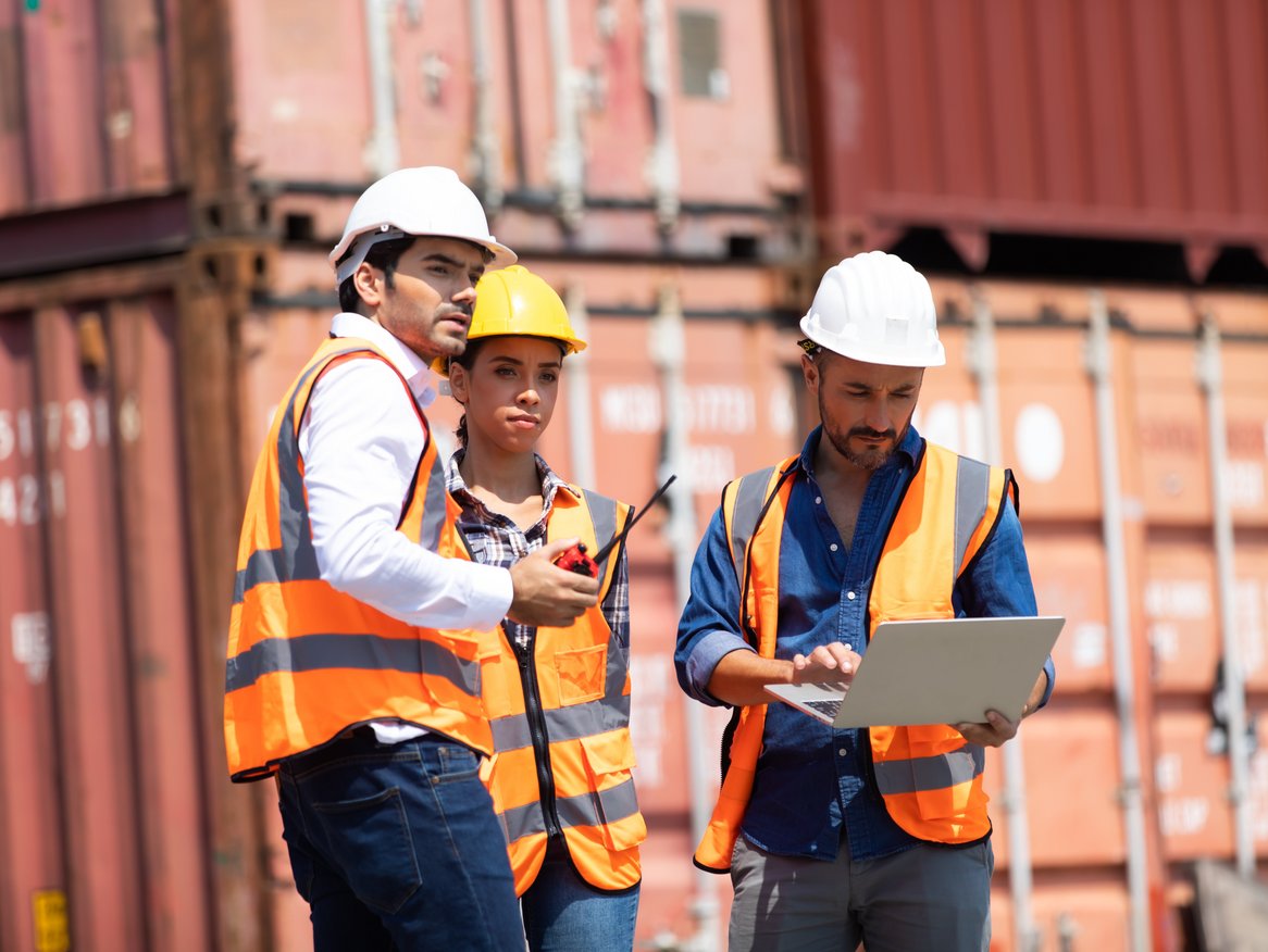 Drei Hafenarbeiter mit Schutzhelmen und Sicherheitswesten besprechen Arbeitsaufgaben mit einem Laptop und Dokumenten vor einem Hintergrund mit gestapelten Frachtcontainern.