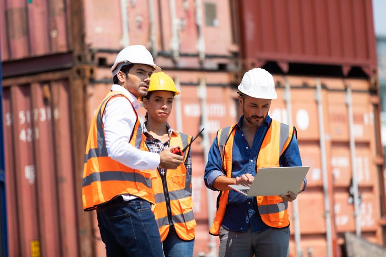 Drei Hafenarbeiter mit Schutzhelmen und Sicherheitswesten besprechen Arbeitsaufgaben mit einem Laptop und Dokumenten vor einem Hintergrund mit gestapelten Frachtcontainern.