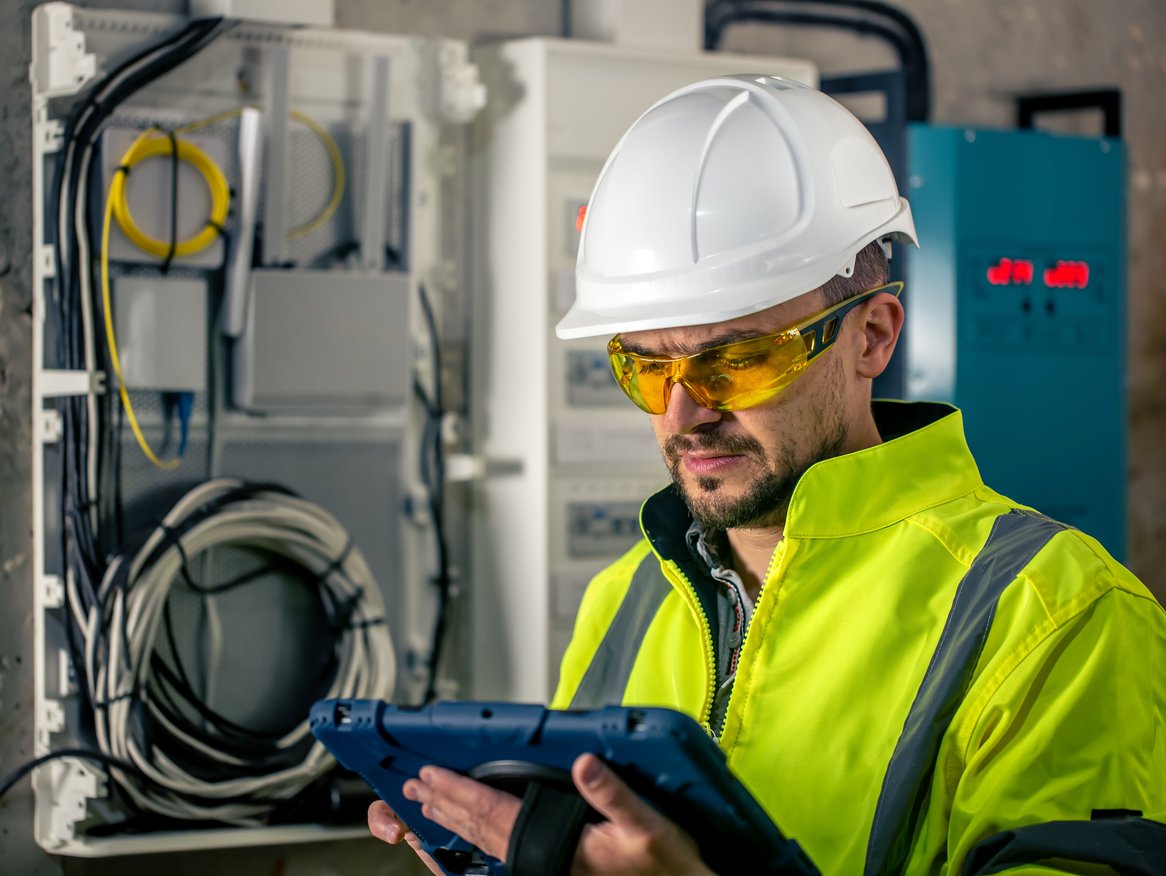 Ein Ingenieur in Schutzausrüstung konzentriert sich auf ein elektronisches Gerät, möglicherweise zur Überwachung oder Steuerung industrieller Prozesse, vor einem Hintergrund mit technischen Installationen.