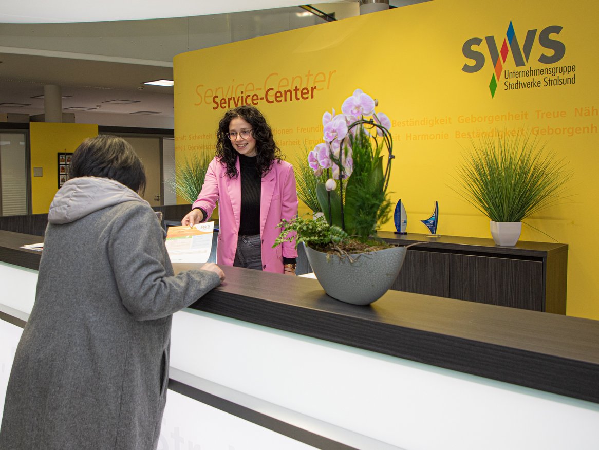 Eine freundliche Angestellte im pinken Mantel bedient eine Kundin am Service-Center der Stadtwerke Stralsund. Im Hintergrund sind Zimmerpflanzen und Auszeichnungen zu sehen sowie die Worte "Vertrauen, Zuverlässigkeit" an der gelben Wand, die die Unternehmenswerte widerspiegeln