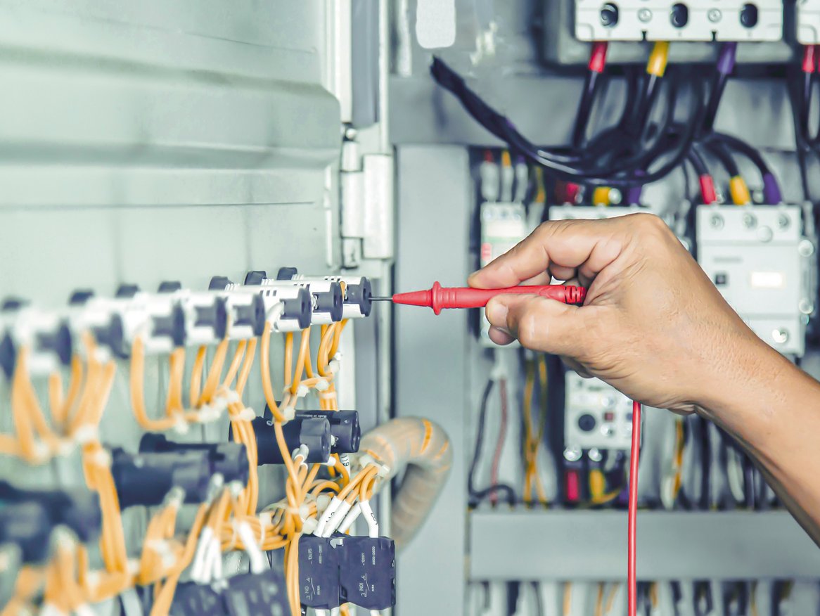 Eine Hand führt mit einem Prüfwerkzeug eine elektrische Messung in einem Schaltschrank durch. Die Komplexität des elektrischen Systems und das Fachwissen, das für solche Arbeiten erforderlich ist, werden durch die gezielte Aktion des Technikers betont.