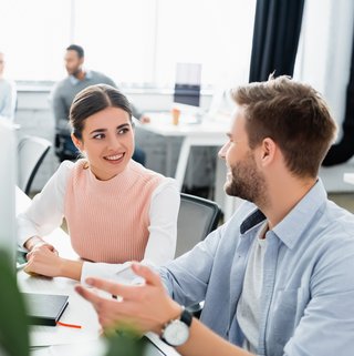 Zwei Kollegen sitzen sich an einem Arbeitsplatz gegenüber und führen ein Gespräch, was auf eine dynamische und kooperative Büroatmosphäre hindeutet. Pflanzen und moderne Computerbildschirme sind ebenfalls sichtbar und unterstreichen das moderne Bürodesign