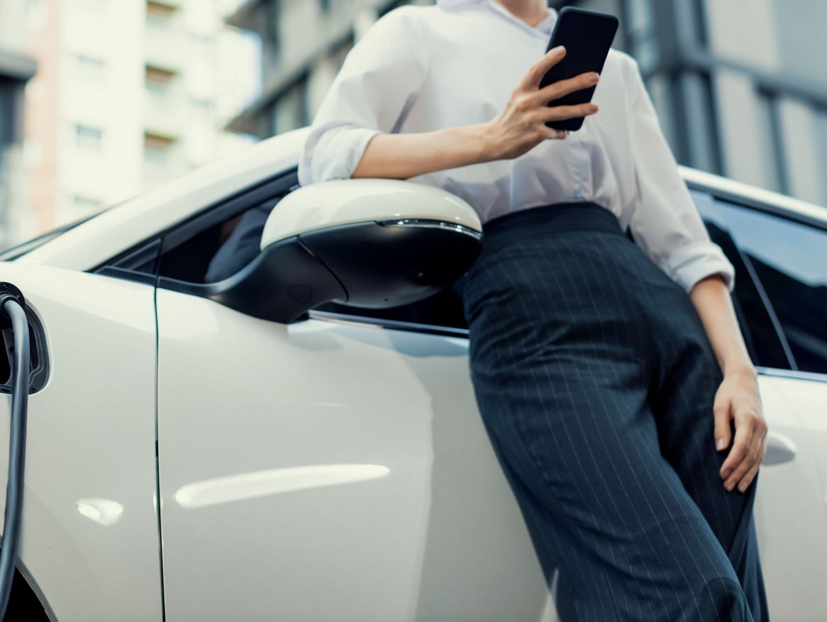Eine Person, nur teilweise im Bild, steht neben einem weißen Elektroauto, das gerade geladen wird, und benutzt ein Smartphone. Die Kleidung lässt auf einen professionellen Kontext schließen, was darauf hindeuten könnte, dass die Person möglicherweise während einer Arbeitspause ihr Auto auflädt.