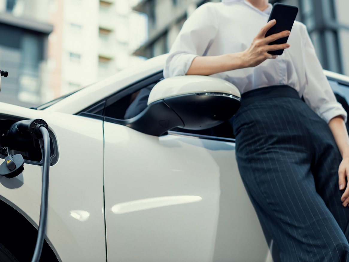 Eine Person, nur teilweise im Bild, steht neben einem weißen Elektroauto, das gerade geladen wird, und benutzt ein Smartphone. Die Kleidung lässt auf einen professionellen Kontext schließen, was darauf hindeuten könnte, dass die Person möglicherweise während einer Arbeitspause ihr Auto auflädt.