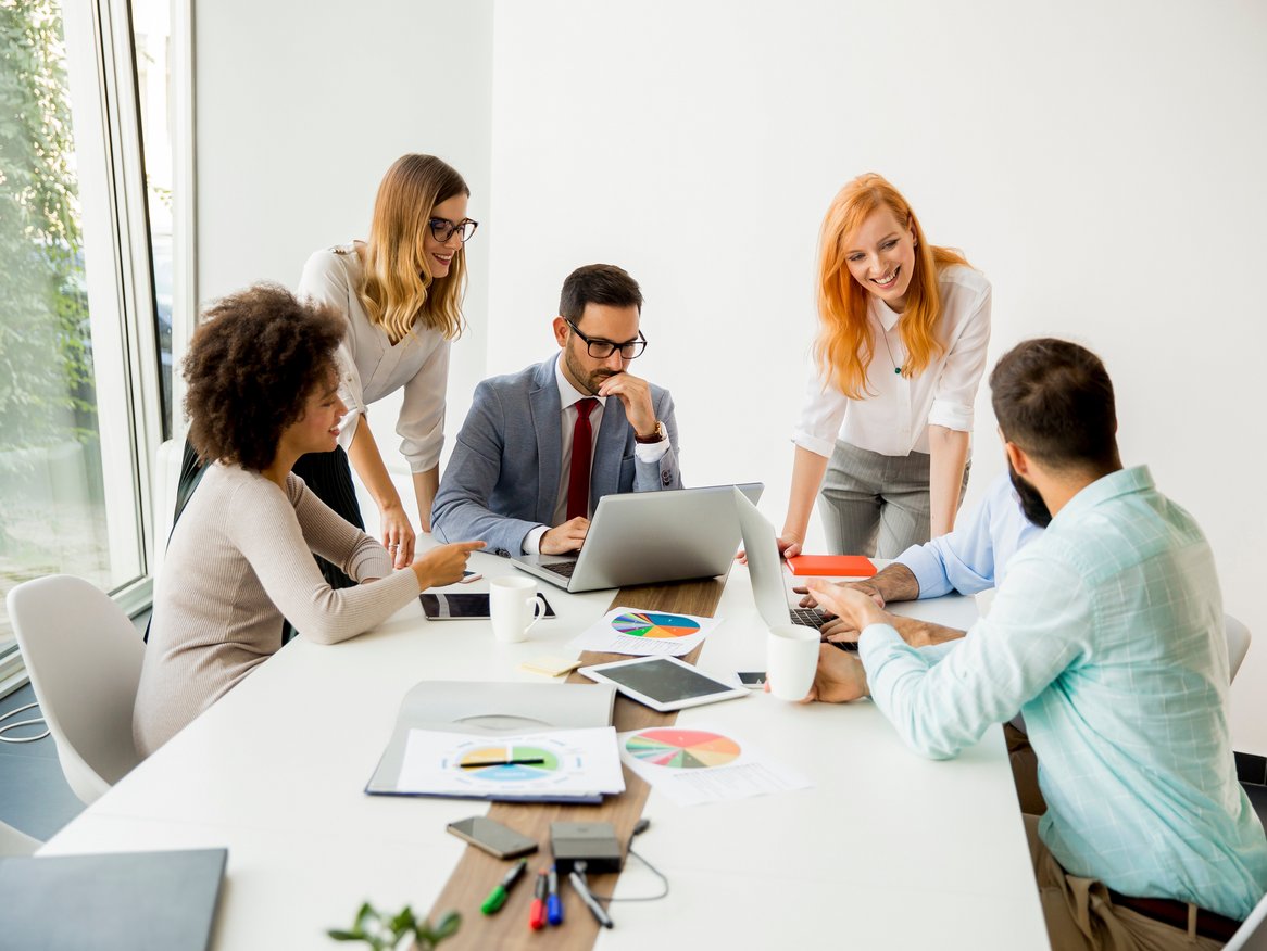 Team von fünf Berufstätigen bei einer Besprechung um einen Tisch mit Laptop, Papieren und Schreibwaren in einem hellen Büro.