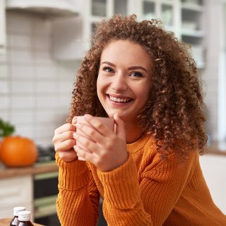 Eine junge Frau mit lockigem Haar und einem warmen, orangefarbenen Pullover hält eine Tasse in den Händen und lächelt herzlich. Ihr Ausdruck und das gemütliche Outfit deuten auf eine entspannte, behagliche Stimmung hin, typisch für eine gemütliche Herbstatmosphäre.