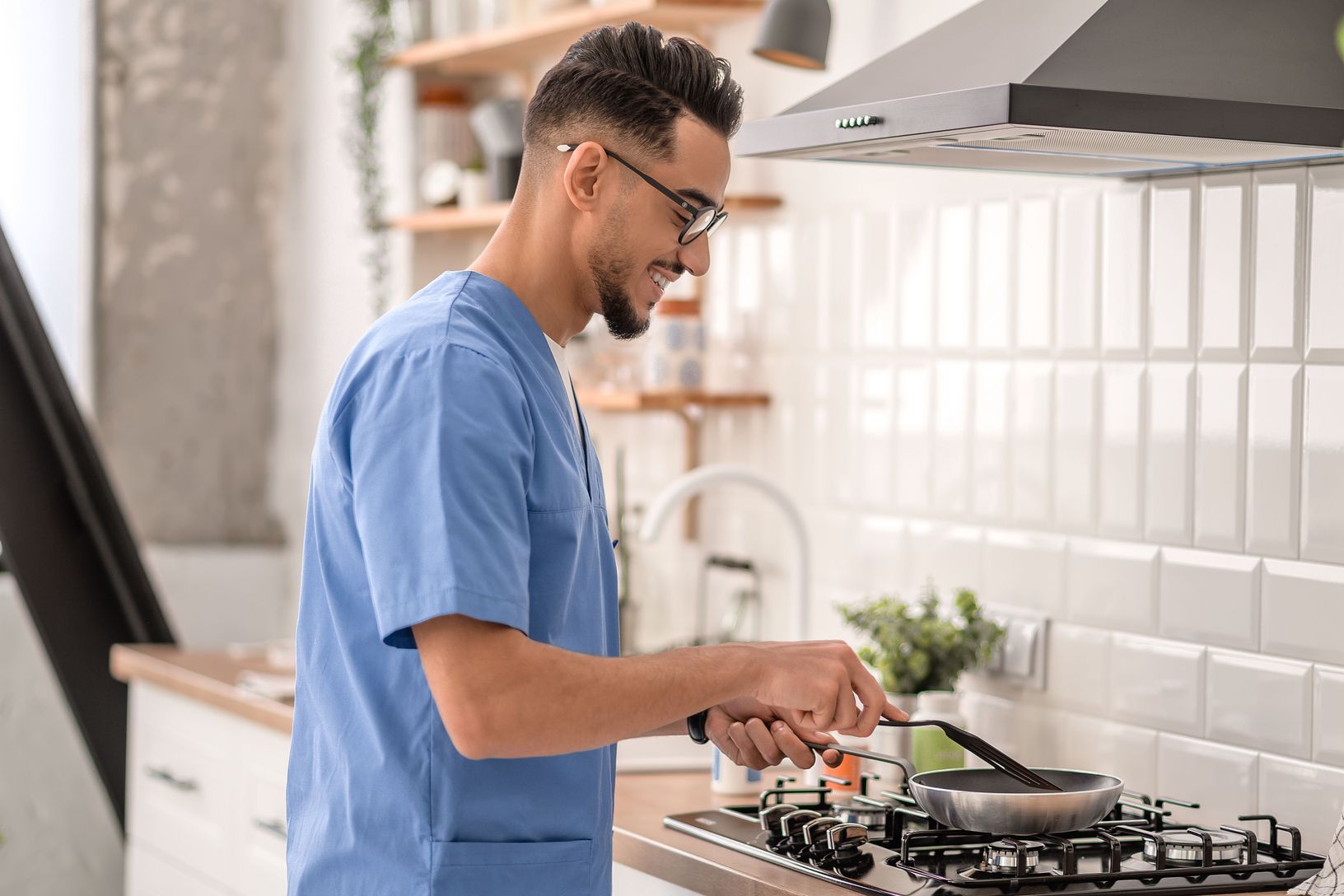 Ein Mann in lässiger Kleidung und mit Brille konzentriert sich auf das Kochen in einer hellen, modern eingerichteten Küche, was den Alltag und die Freuden des Zubereitens von Mahlzeiten zu Hause darstellt.