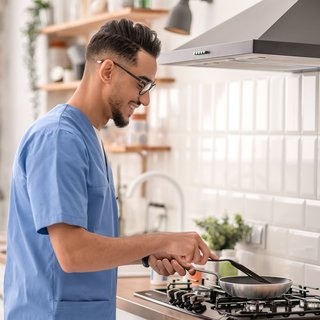 Ein Mann in lässiger Kleidung und mit Brille konzentriert sich auf das Kochen in einer hellen, modern eingerichteten Küche, was den Alltag und die Freuden des Zubereitens von Mahlzeiten zu Hause darstellt.