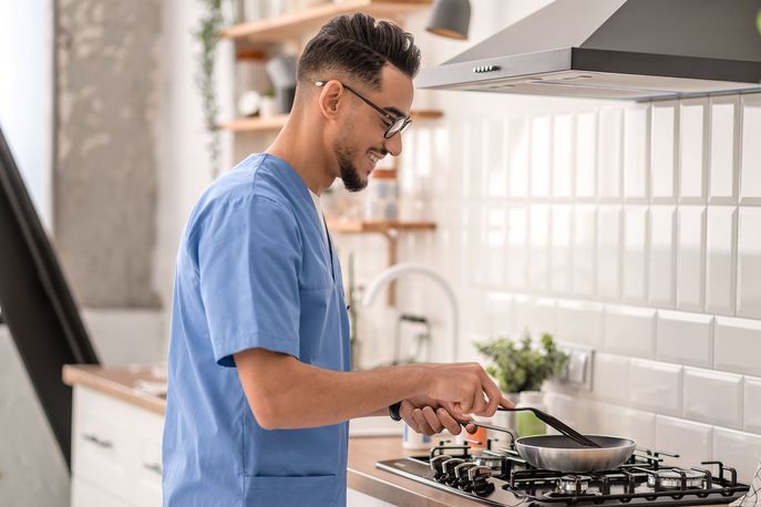 Ein Mann in lässiger Kleidung und mit Brille konzentriert sich auf das Kochen in einer hellen, modern eingerichteten Küche, was den Alltag und die Freuden des Zubereitens von Mahlzeiten zu Hause darstellt.