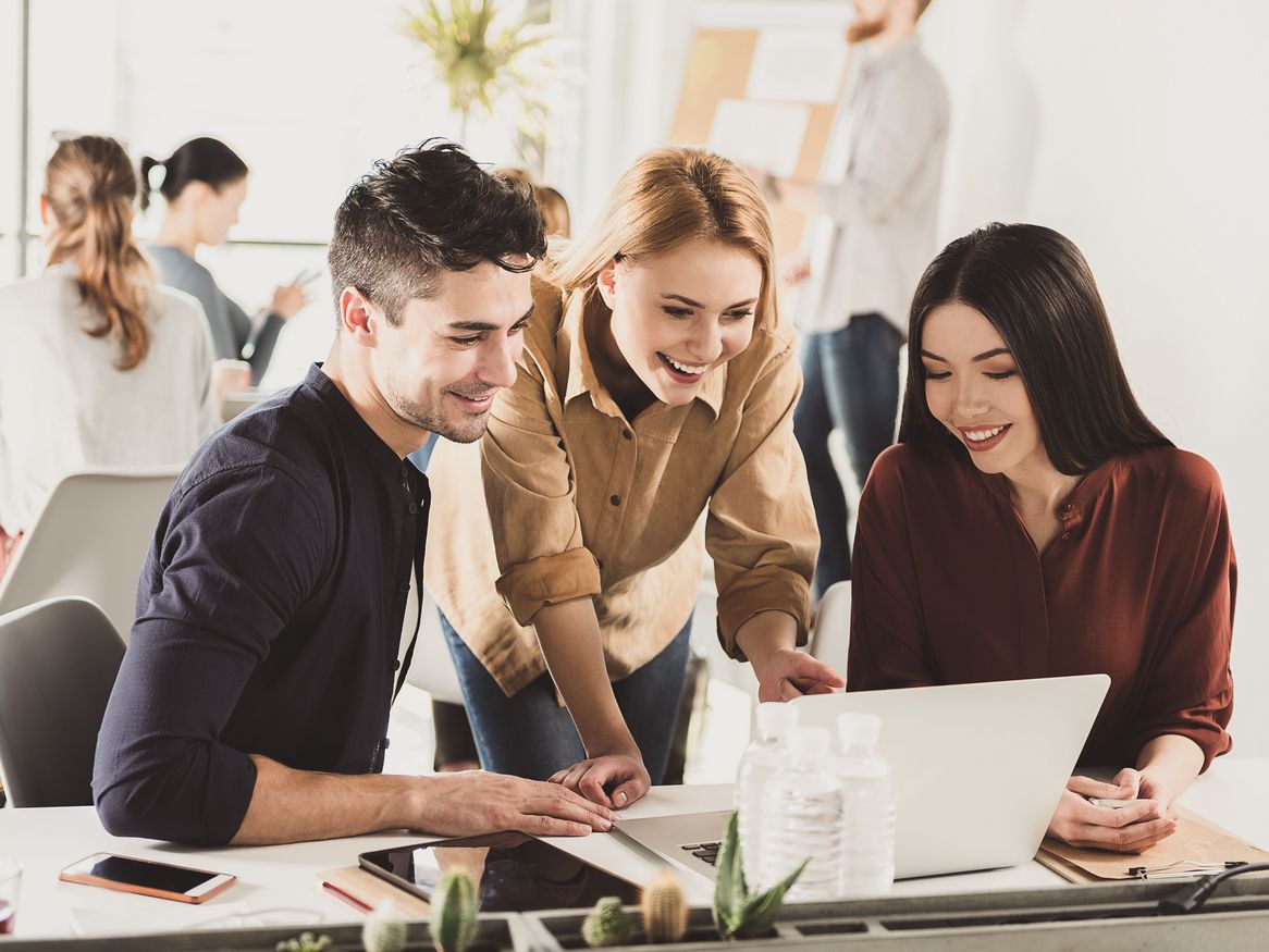 Drei junge Berufstätige, zwei Frauen und ein Mann, führen eine gemeinsame Diskussion über ein Dokument, mit weiteren Kollegen, die im Hintergrund arbeiten.
