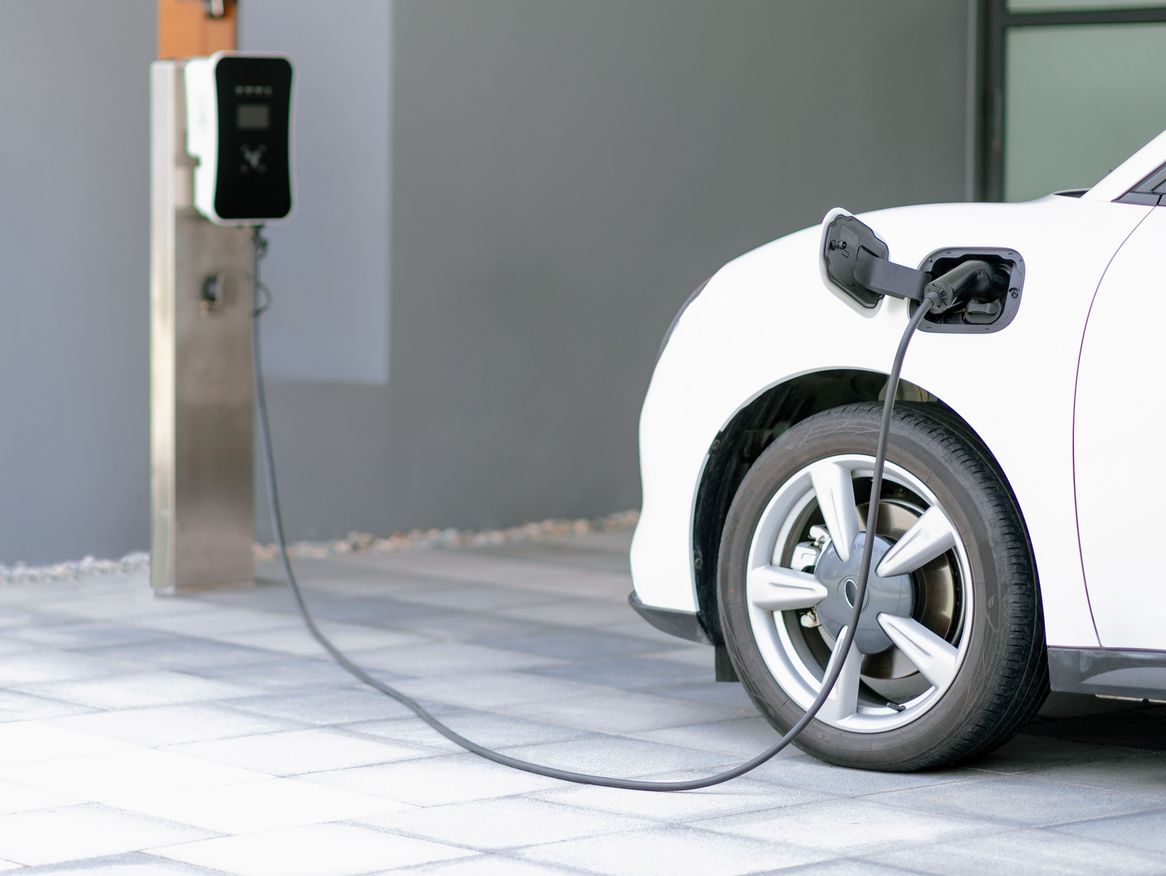 Weißes Elektroauto beim Aufladen an einer öffentlichen Ladesäule mit einem angeschlossenen Ladekabel, vor einer modernen, grauen Gebäudewand, ein Bild der zunehmenden Elektromobilität.