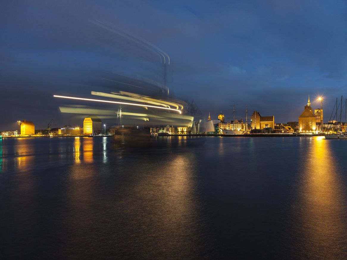 Eine Hafenansicht bei Nacht mit langzeitbelichteten Lichtern, die sich über das Wasser bewegen, was einen dynamischen Effekt erzeugt. Die historischen Gebäude im Hintergrund bieten einen schönen Kontrast zu den modernen Lichtspuren.
