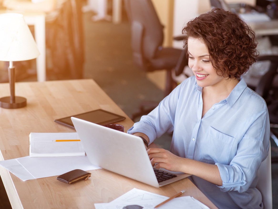 Eine fröhliche Frau mit lockigem Haar arbeitet konzentriert an einem Laptop, umgeben von Papieren und einem Smartphone, in einem hell beleuchteten Büro.