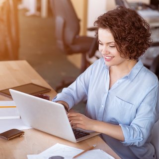 Eine fröhliche Frau mit lockigem Haar arbeitet konzentriert an einem Laptop, umgeben von Papieren und einem Smartphone, in einem hell beleuchteten Büro.