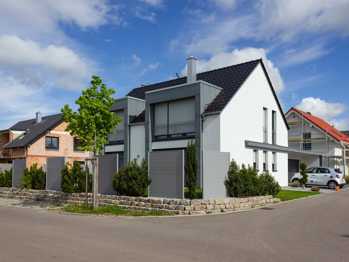 Moderne Wohnhäuser mit sauberen Linien, Giebeldächern und gepflegten Vorgärten in einer ruhigen Vorstadtsiedlung an einem sonnigen Tag mit einigen Wolken am blauen Himmel.