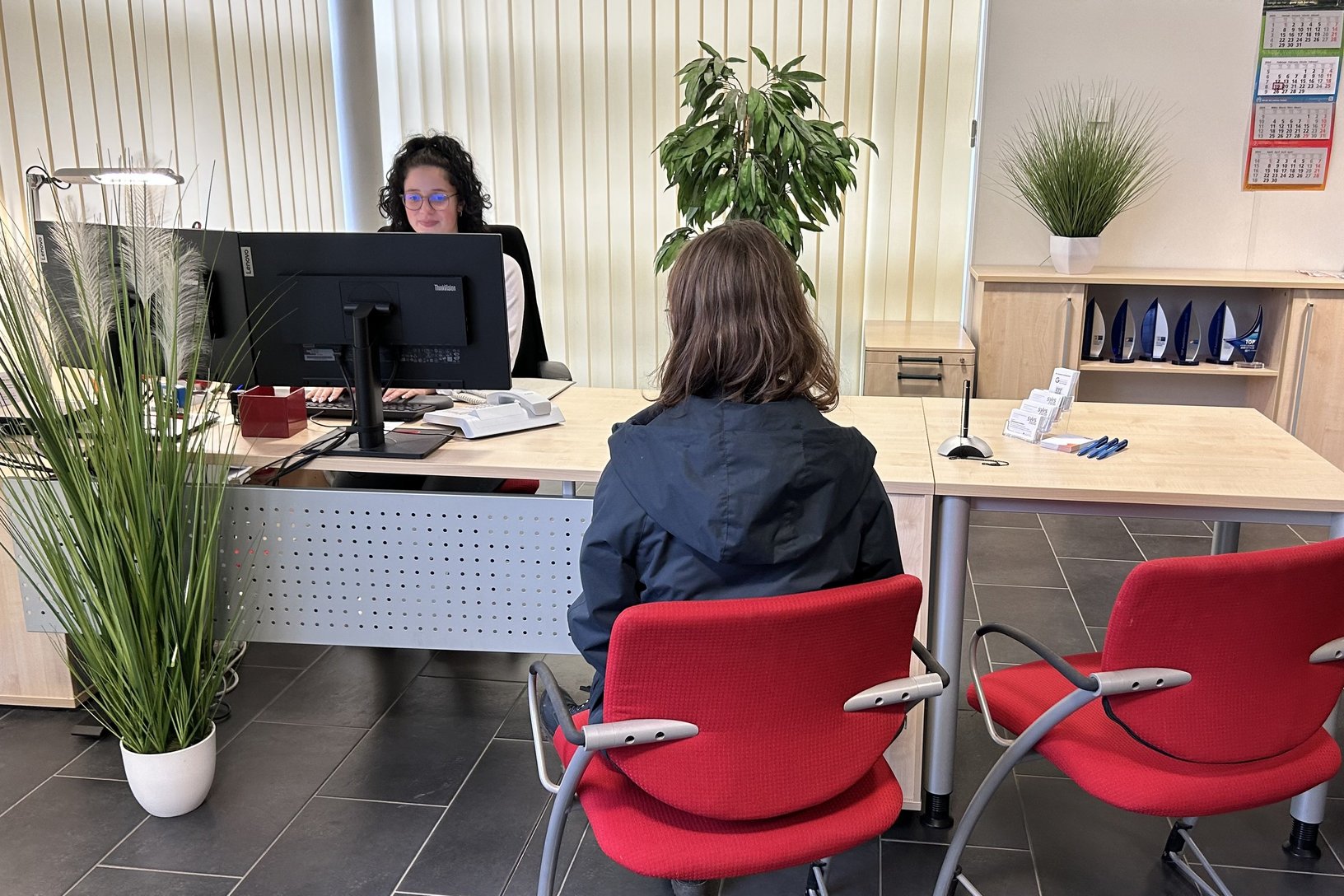 Eine Person wartet im Vordergrund auf einem roten Stuhl in einem Büro, während eine Angestellte hinter einem Computerbildschirm arbeitet, umgeben von Pflanzen und Büromaterialien.