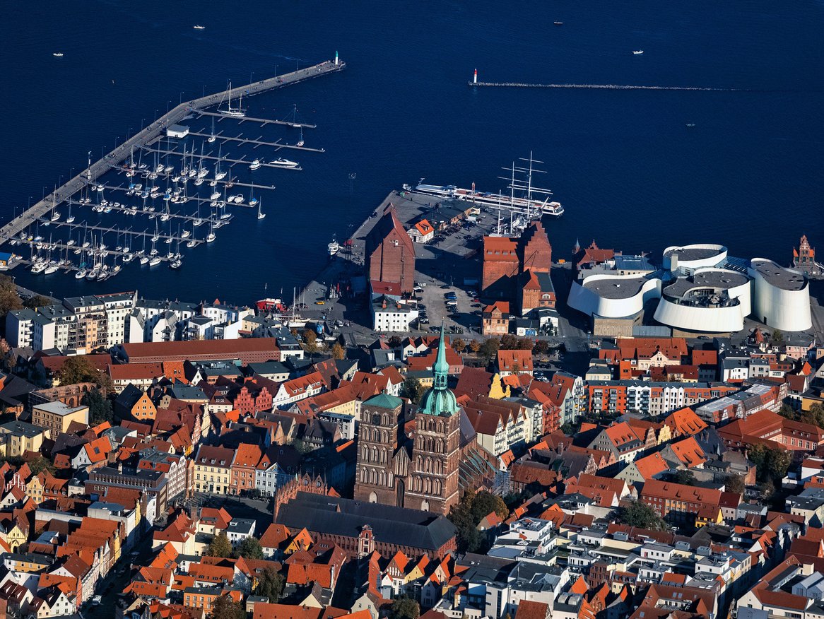 Luftaufnahme des Hafens von Stralsund, die eine organisierte Anordnung von Segelbooten am Kai zeigt, mit dem markanten Ozeaneum und historischen Gebäuden im Vordergrund, die sich gegen das glänzende blaue Wasser der Ostsee abheben.