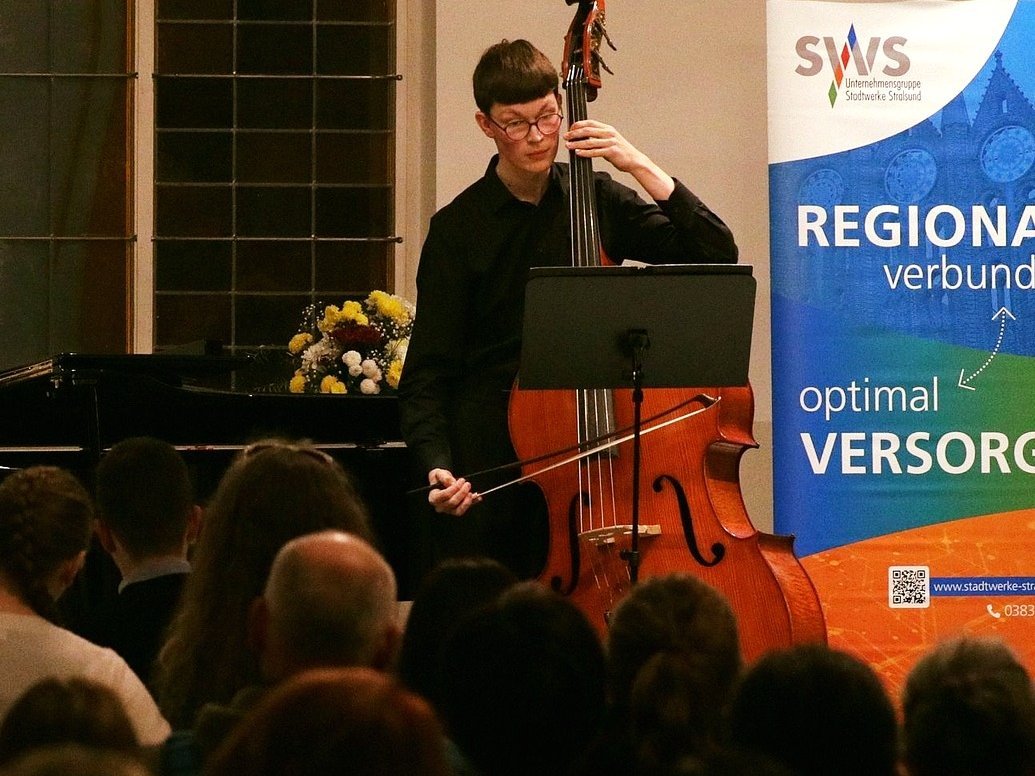 Ein Cellist spielt leidenschaftlich vor einem Publikum, im Hintergrund ist ein Banner mit der Aufschrift "REGIONAL... VERSORGUNG" zu sehen