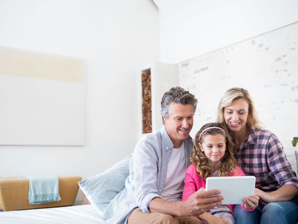 Eine Familie mit einem Kind sitzt gemeinsam auf dem Bett und betrachtet ein Tablet. Sie lächeln und genießen eine gemütliche Zeit in einem hellen, modern eingerichteten Schlafzimmer.