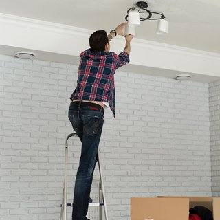 Ein Mann auf einer Leiter hält eine Überwachungskamera, während eine Frau ihm Anweisungen gibt. Dieses Bild zeigt die Installation von Sicherheitstechnik in einem modernen Wohnraum, was auf die Bedeutung von Sicherheit im Eigenheim hinweist.