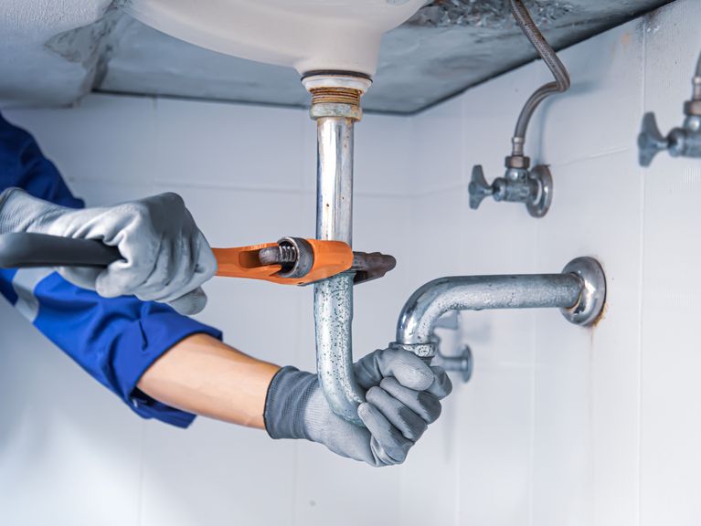 Hände eines Klempners in Handschuhen, der mit einer Rohrzange an der Verrohrung unter einem Waschbecken arbeitet, mit sichtbaren Wasserhähnen im Hintergrund.