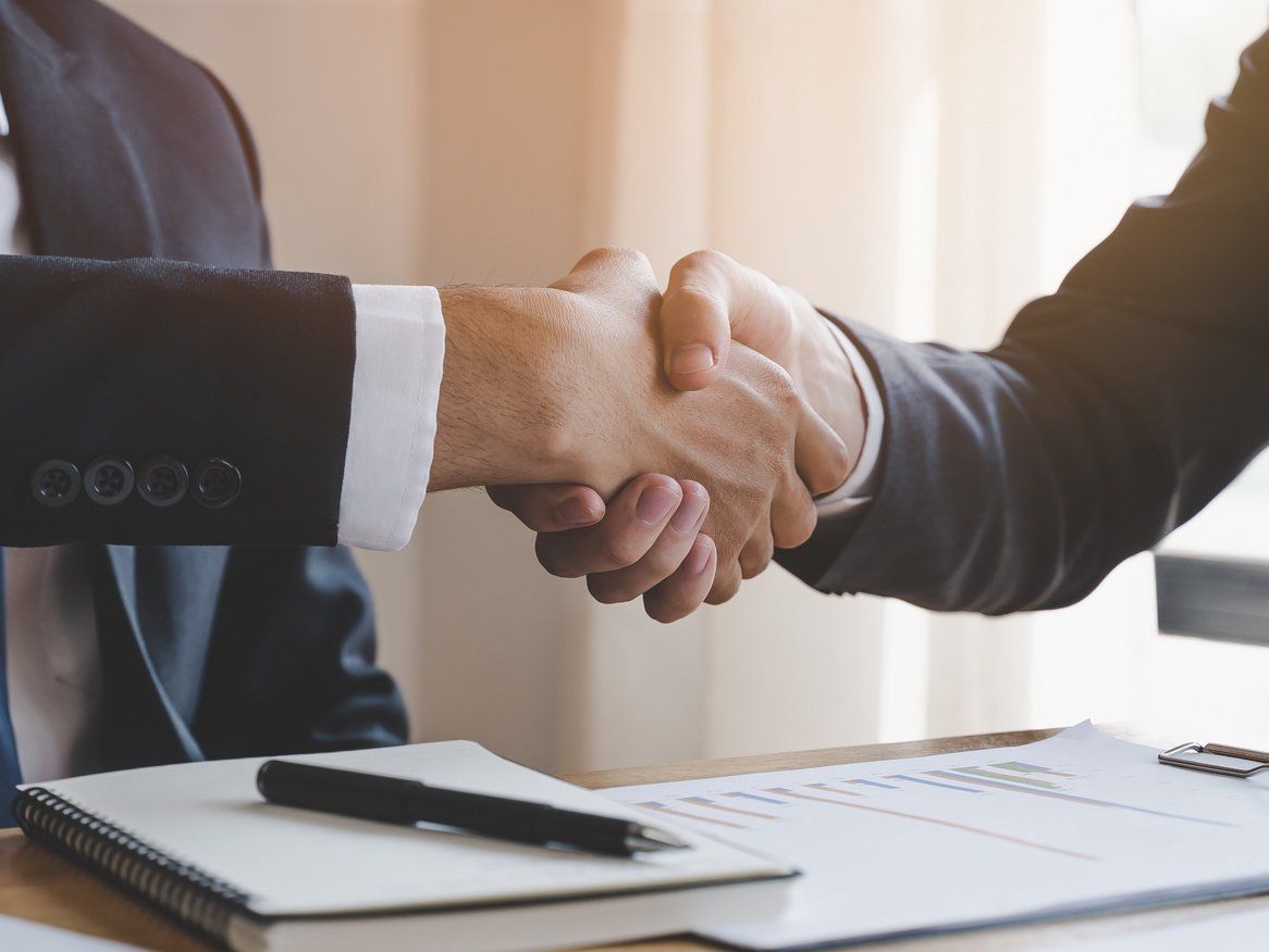 Zwei Geschäftsleute bei einem Handschlag über einem Tisch, der mit Dokumenten bedeckt ist, ein klassisches Bild für erfolgreiche Verhandlungen und Partnerschaften im Geschäftsumfeld.