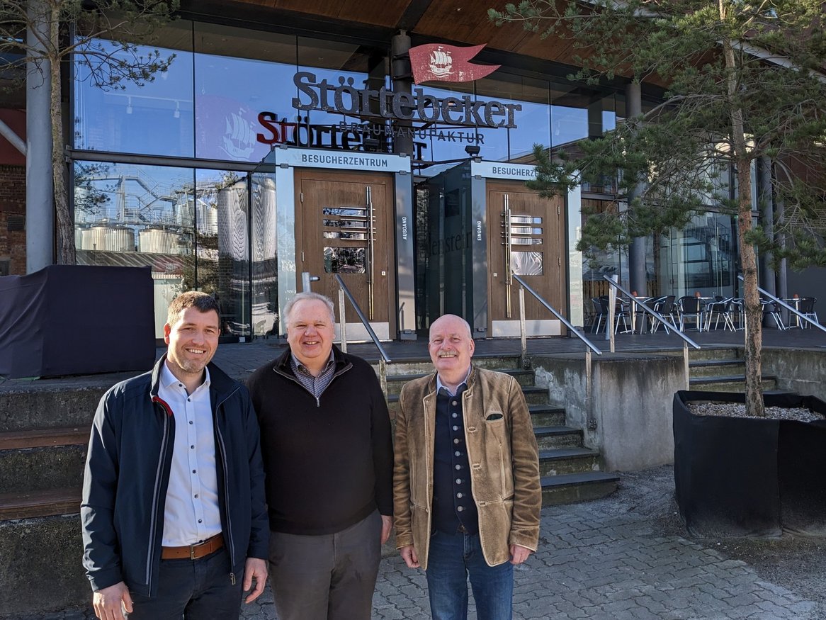 Drei lächelnde Personen stehen vor dem Eingang der Störtebeker Braumanufaktur, erkennbar an dem großen Schild über den Glastüren, umgeben von Pflanzkübeln und unter einem strahlend blauen Himmel.