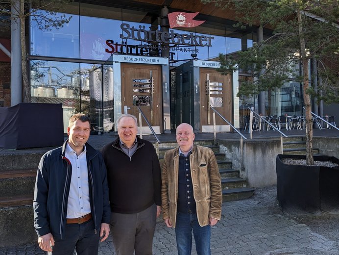 Drei lächelnde Personen stehen vor dem Eingang der Störtebeker Braumanufaktur, erkennbar an dem großen Schild über den Glastüren, umgeben von Pflanzkübeln und unter einem strahlend blauen Himmel.